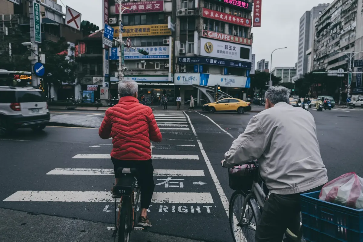 Elderly men riding bicycles across a pedestrian lane in a bustling city in Taiwan, showcasing urban life.