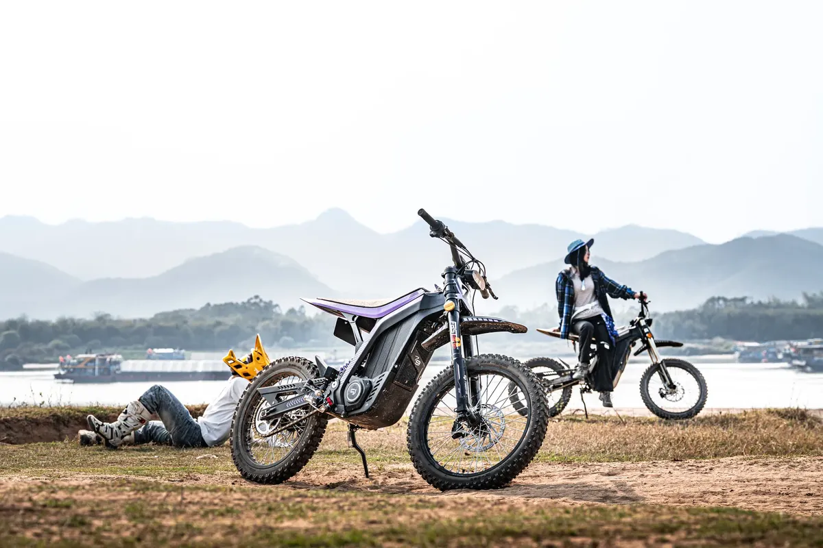 Two individuals on electric dirt bikes near a serene lake with mountains in the background.
