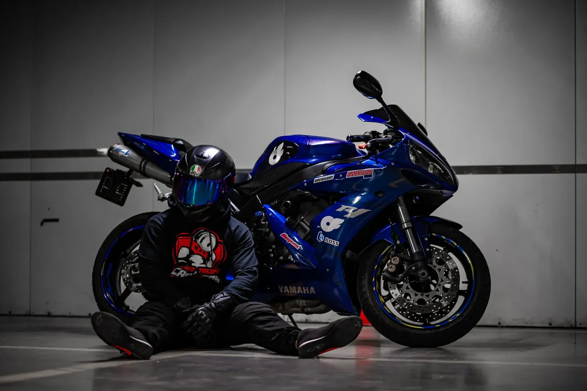 A motorcycle rider in black gear sits by a blue sports bike indoors, showing urban style.