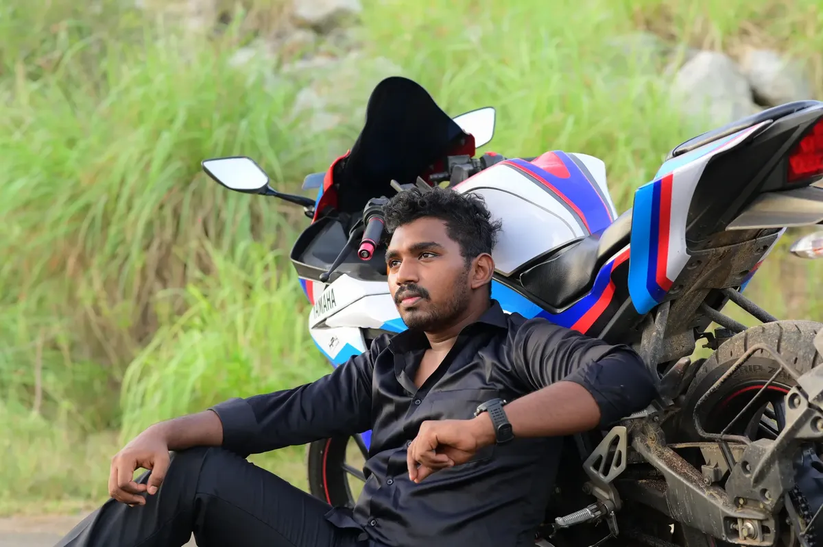 A man in a black shirt sitting pensively near his Yamaha motorcycle in a rural setting.